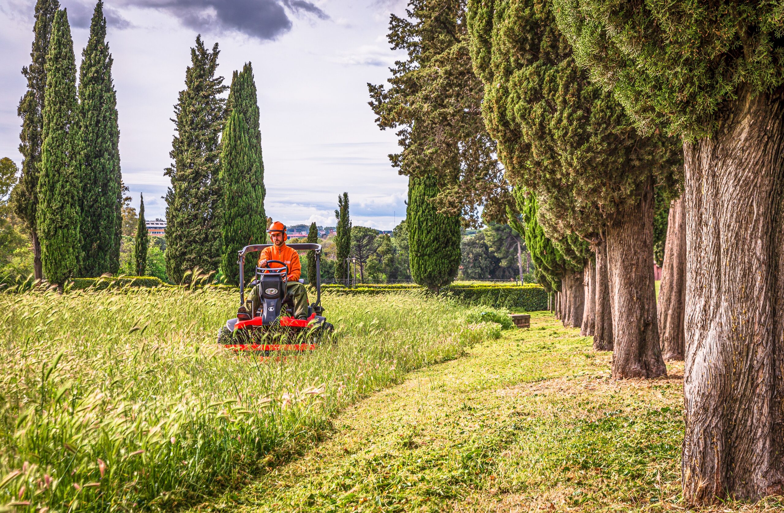A man operates a lawn mower in a picturesque Italian landscape with cypress trees.